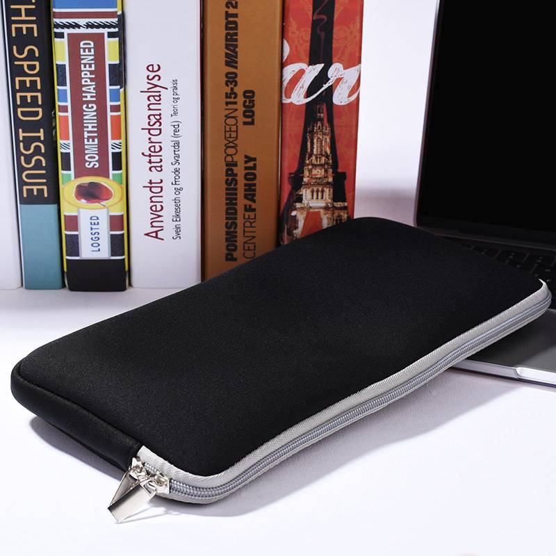Schwarze Laptop-Schutzhülle mit Reißverschluss auf einem weißen Schreibtisch neben einem Laptop, mit Büchern, die im Hintergrund stehen.