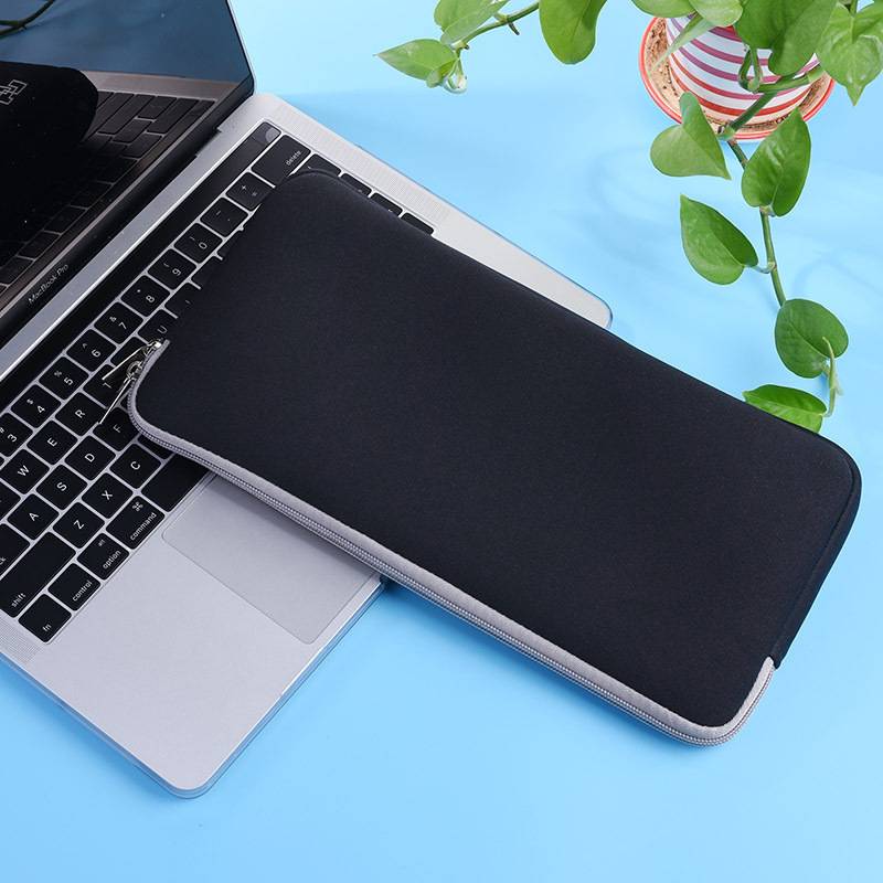 Schwarze reißverschlossene Laptophülle, die diagonal auf einem aufgeklappten silbernen Laptop vor einem blauen Hintergrund mit einer Topfpflanze liegt.
