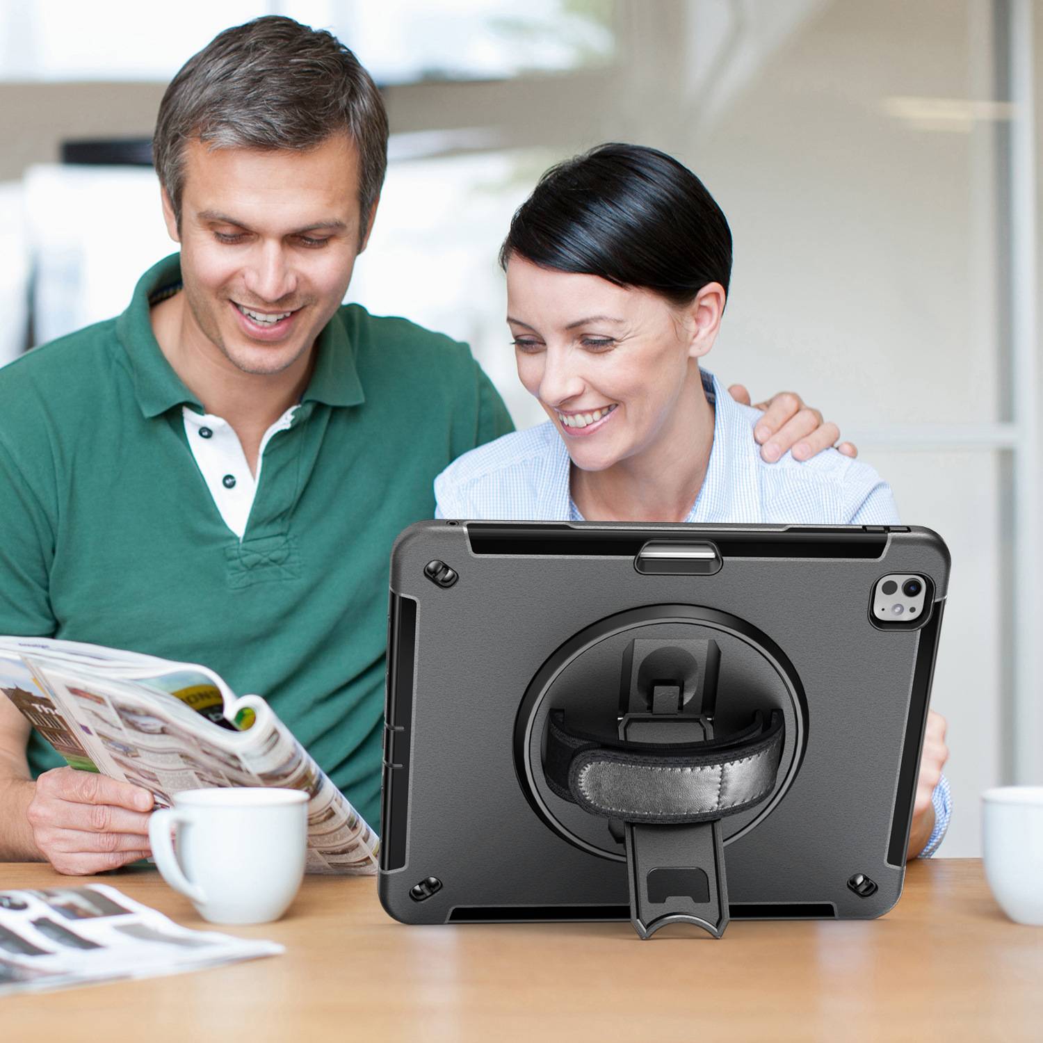 Ein lächelndes Paar sitzt an einem Tisch und schaut auf ein Tablet, während eine Person eine Zeitung liest und Kaffeetassen in der Nähe stehen.