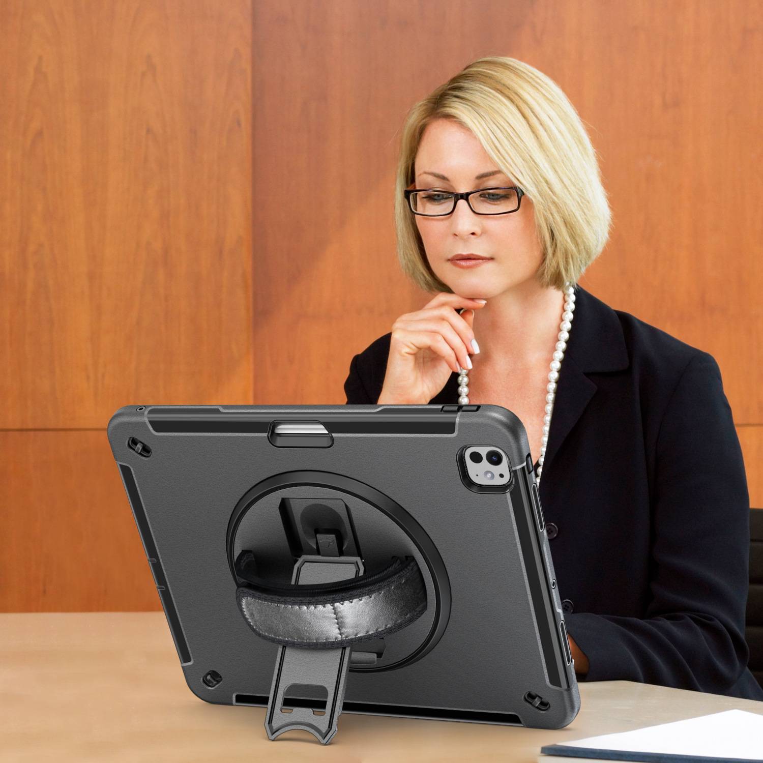 Blonde Frau mit Brille und Business-Kleidung studiert ein Tablet auf einem Ständer an einem Tisch, die Hand am Kinn, vor einer Holzpaneelwand.