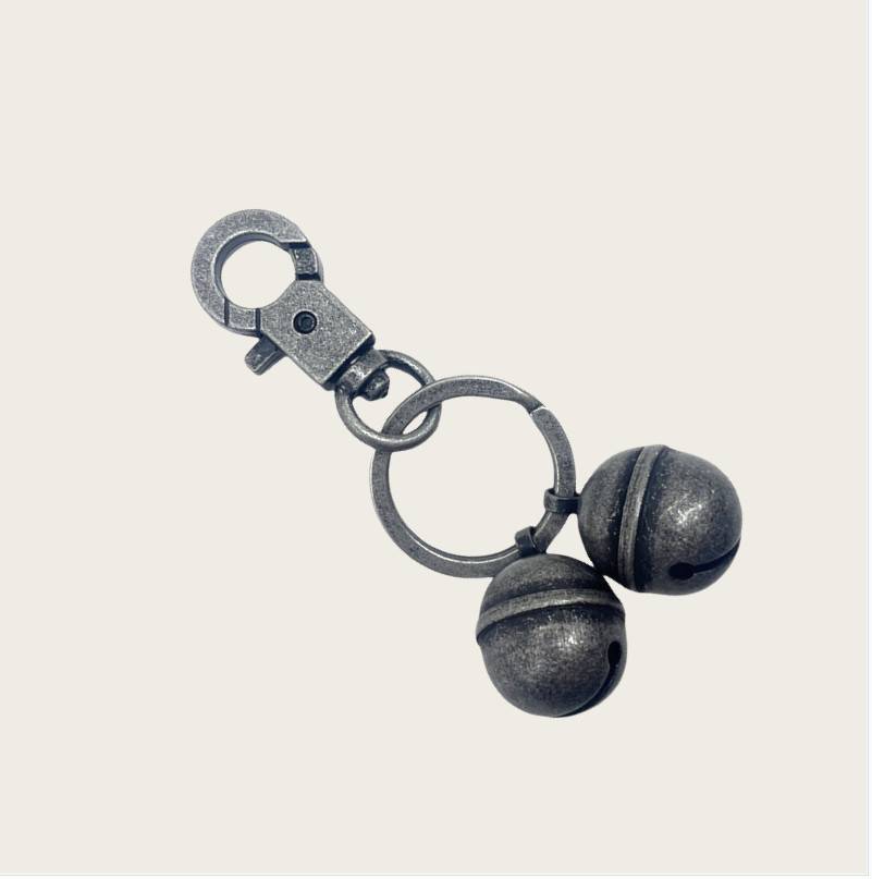 Metallschlüsselanhänger mit zwei dunklen glockenförmigen Anhängern, die durch Ringe an einem einfachen, hellen Hintergrund befestigt sind.