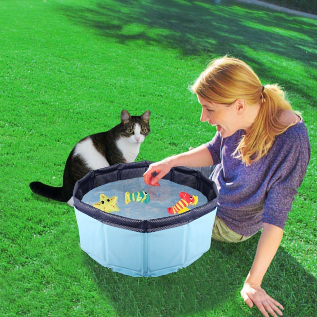 Eine Frau kniet auf dem Gras und spielt mit einer Katze mit Hilfe eines tragbaren Wassersspielzeugs in Form eines Pools mit schwimmenden Fischspielzeugen.