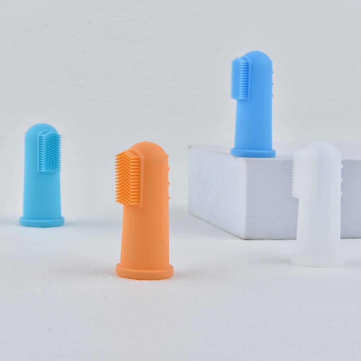 Drei bunte Silikon-Fingerzahnbürsten auf einem einfachen weißen Hintergrund, zwei stehen auf weißen Blöcken und eine steht allein.