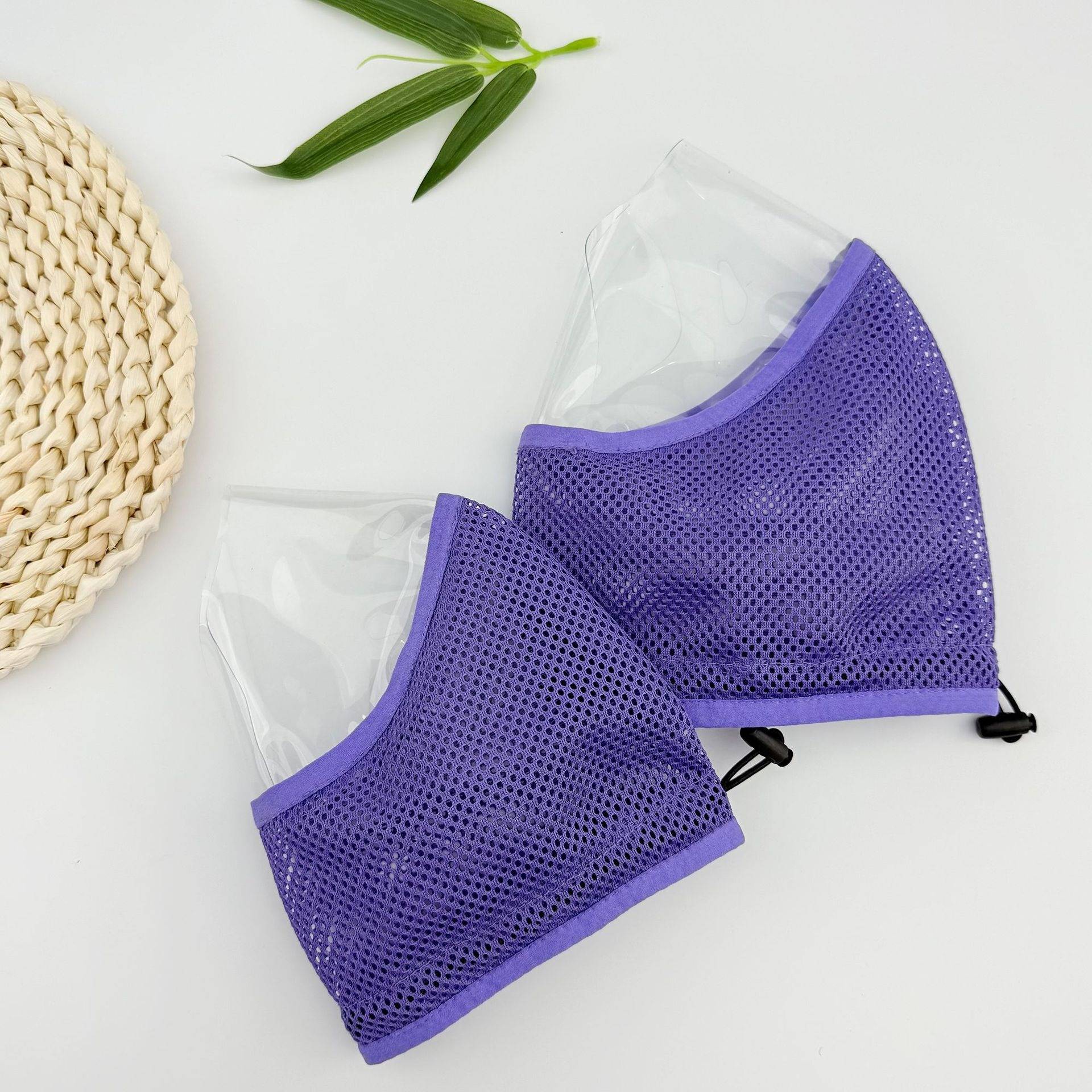 Zwei violette Mesh-Gesichtsmasken mit transparenten Kunststoffplatten, auf einer weißen Oberfläche neben einer geflochtenen Matte und grünen Blättern angeordnet.