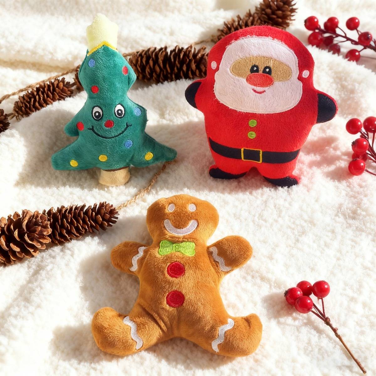 Drei festliche Plüschtiere auf einer weißen Decke: ein Weihnachtsbaum, der Weihnachtsmann und ein Lebkuchenmann, mit Tannenzapfen und roten Beeren um sie herum.