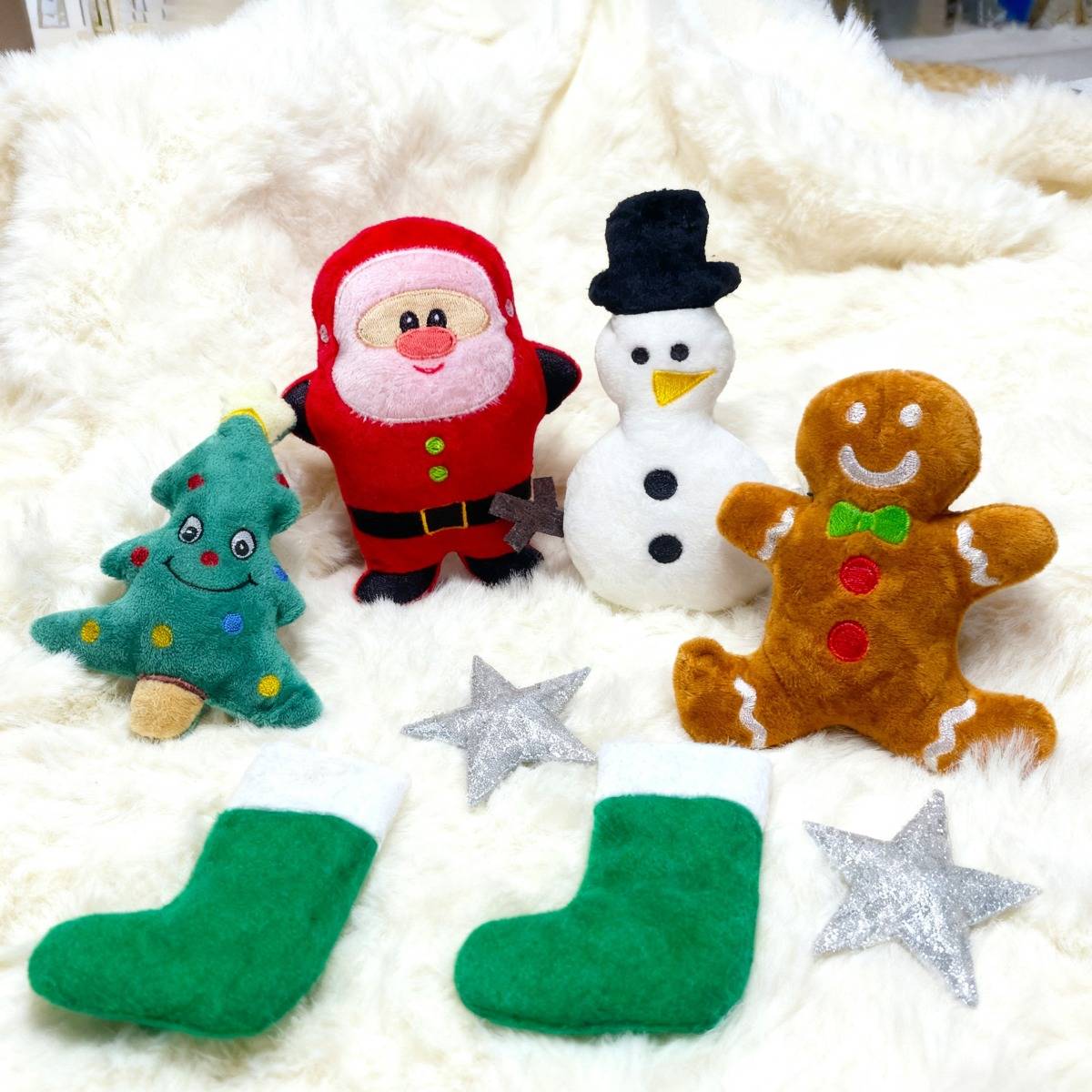 Plüsch-Weihnachtsfiguren auf einer weißen Decke: ein Baum, ein Weihnachtsmann, ein Schneemann, ein Lebkuchenmann und Strumpf-Ornamente mit silbernen Sternen.