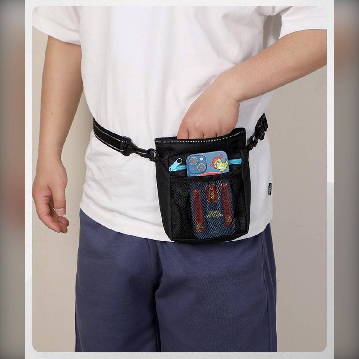 Person mit einer schwarzen Hüfttasche, die in die vordere Tasche greift, während sie vor einem einfachen Hintergrund steht.