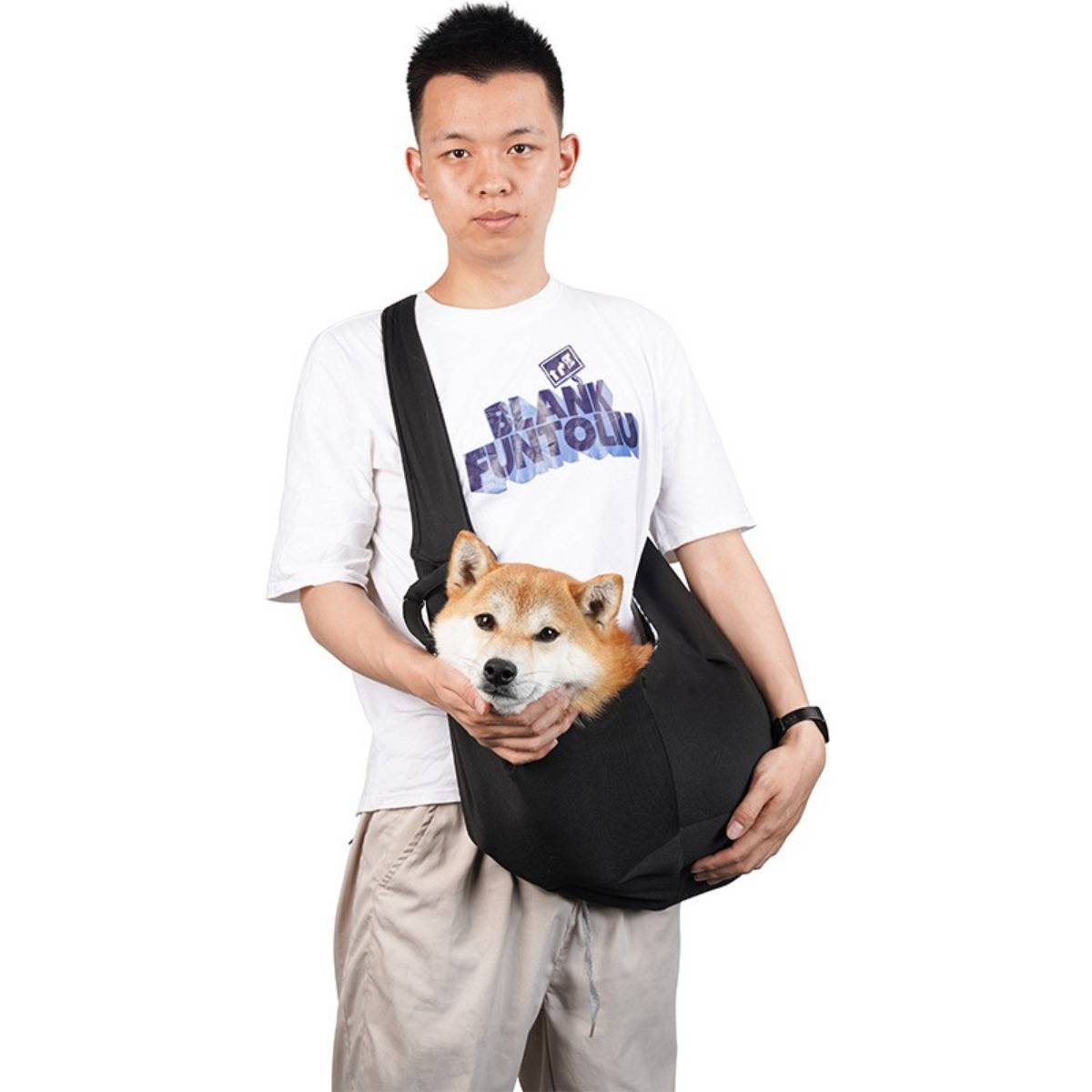 Junger Mann in weißem T-Shirt trägt einen Shiba Inu in einer schwarzen Umhängetasche vor einem einfachen weißen Hintergrund.
