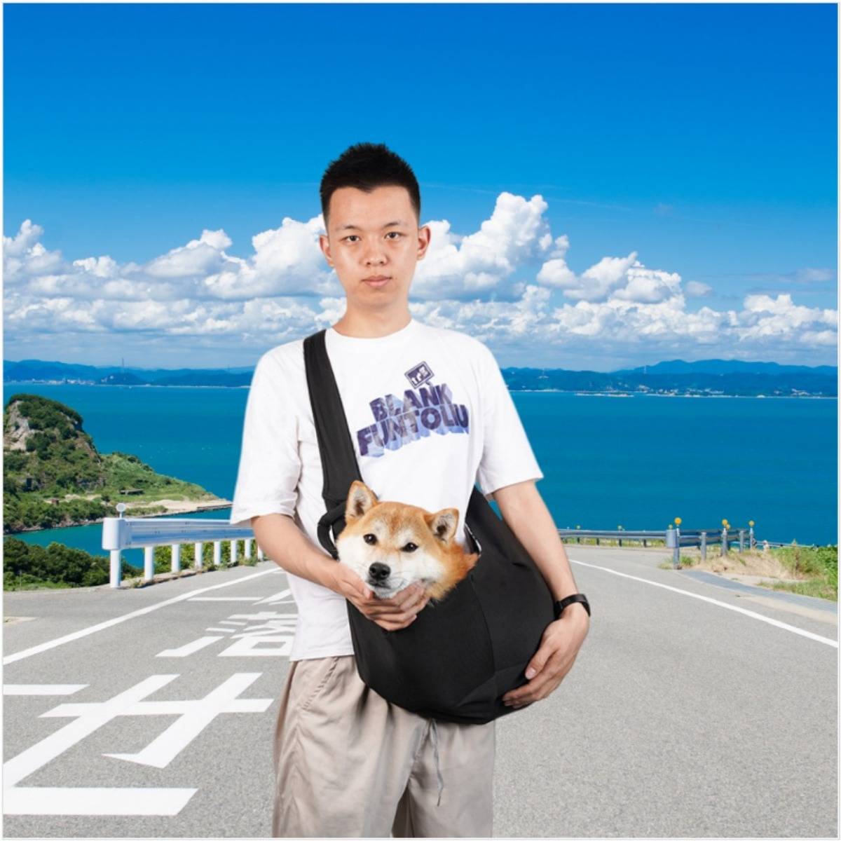 Ein junger Mann steht auf einer Küstenstraße unter einem blauen Himmel und hält einen Shiba Inu in einer schwarzen Tragetasche, mit dem Meer und Bergen hinter ihm.