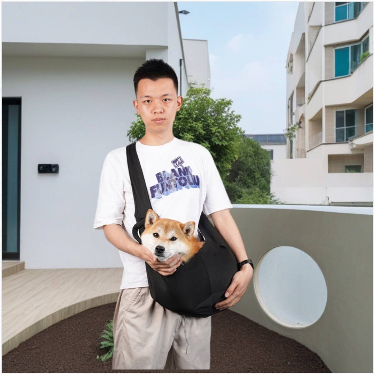Ein junger Mann steht draußen und hält einen Shiba Inu in einer schwarzen Fronttrage zwischen Wohnhäusern.