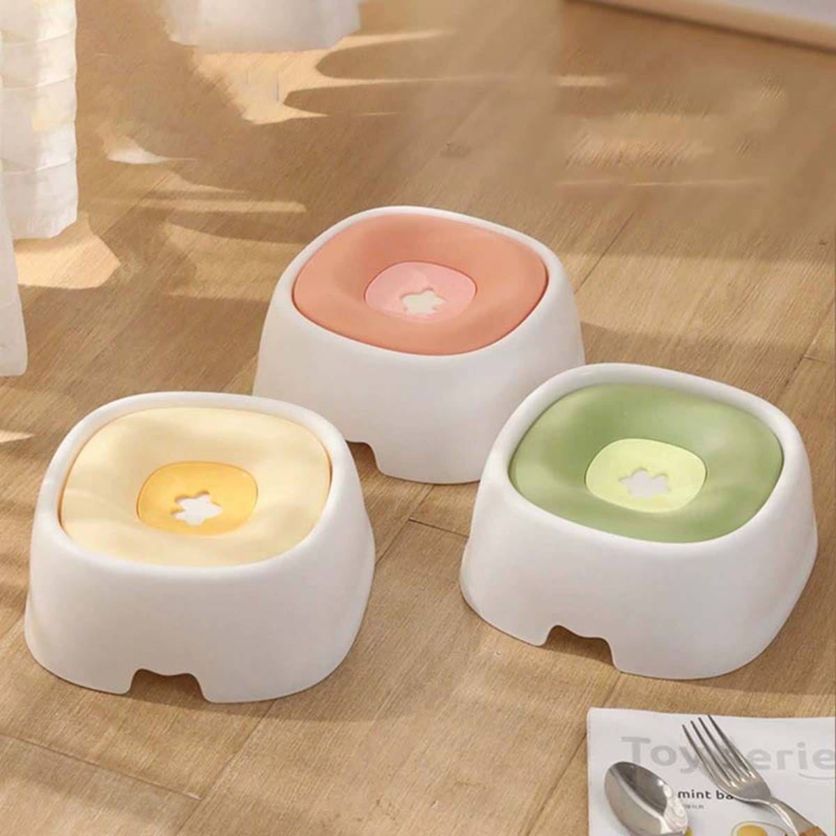 Drei kleine weiße Haustierhocker mit Pastell-Kissen – gelb, rosa und grün – stehen auf einem Holzboden in einem hellen Raum.