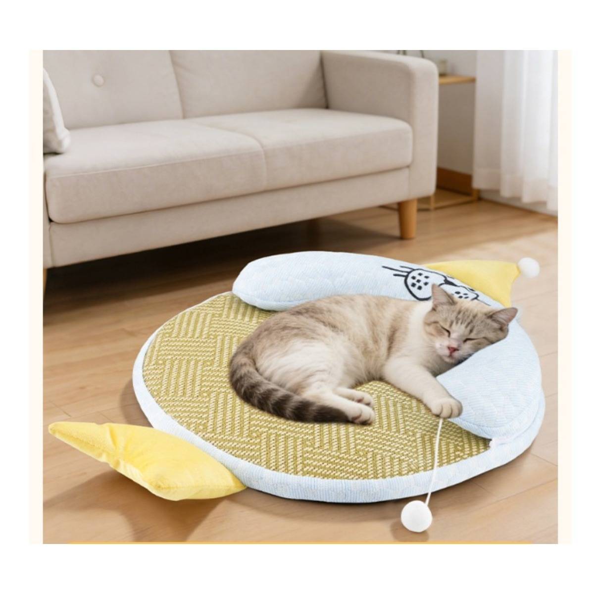 Eine schlafende Katze ruht auf einem runden, gepolsterten Katzenbett mit einem Spielball in einem hellen Wohnzimmer neben einem Sofa.