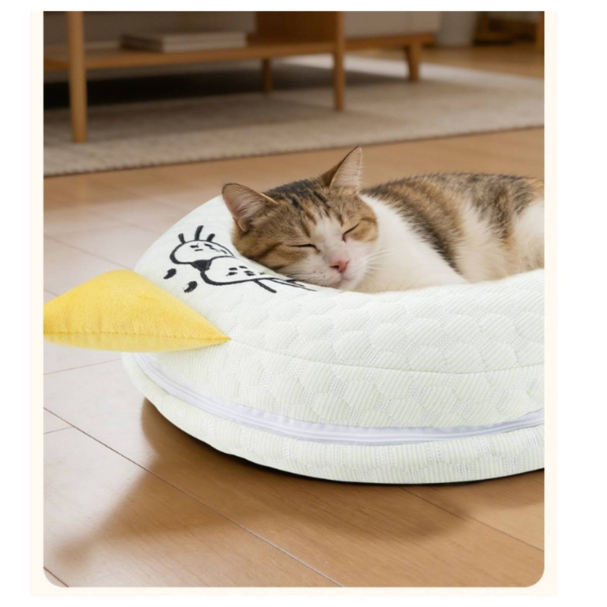 Eine schlafende getigerte Katze ruht auf einem weißen runden Kissen in Form einer gelb akzentuierten Ente auf einem Holzboden in einem Wohnungsinnern.