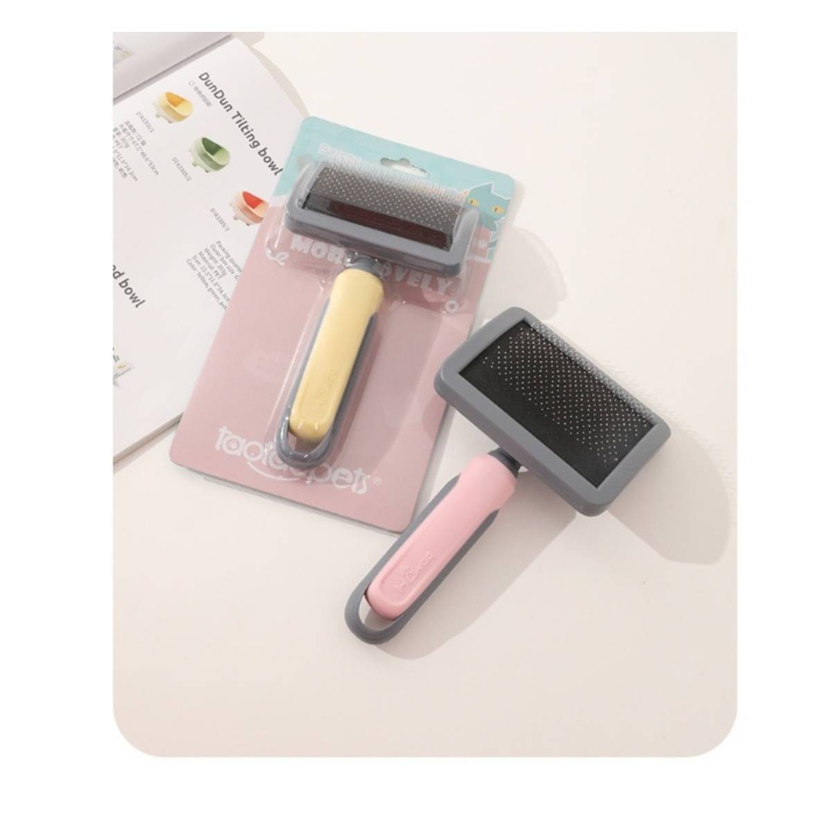Zwei Haustierkardätschen in Verpackung, eine mit gelbem Griff und eine mit rosa Griff, auf einer hellen Oberfläche mit einem Teil eines Produktinformationsblatts gezeigt.