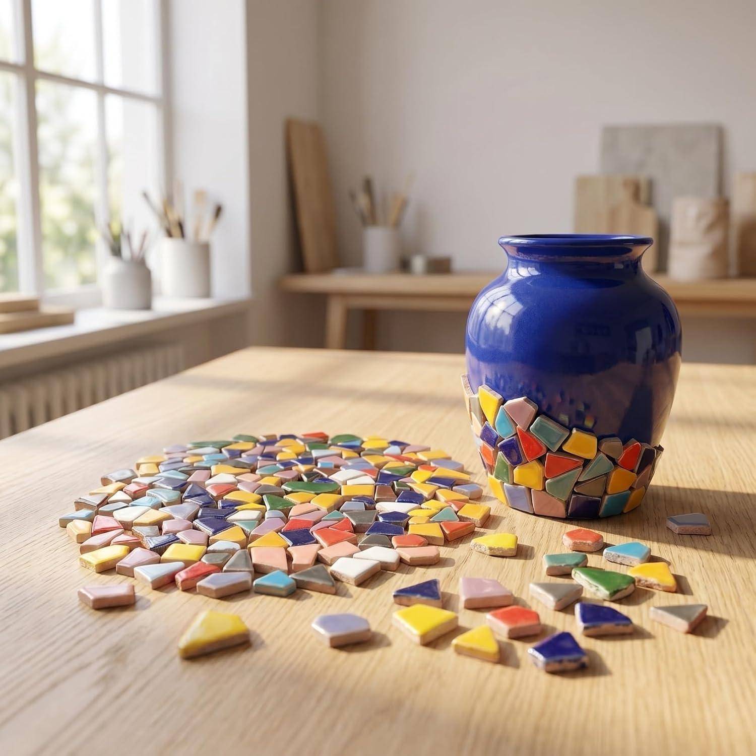 Blaue Keramikvase, die mit bunten Mosaikfliesen auf einem Holztisch in einem hellen Studio dekoriert wird.