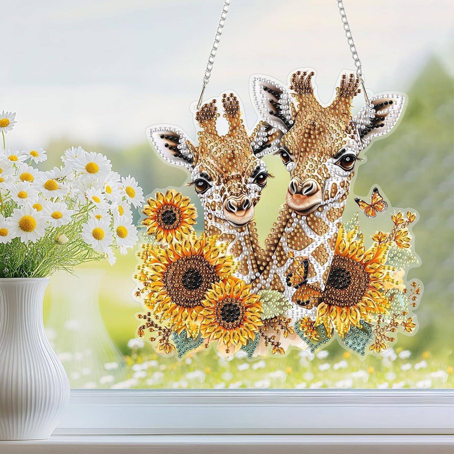 Zwei geschmückte Giraffen-Ornamente hängen an Ketten, eingerahmt von Sonnenblumen und Schmetterlingen neben einer Vase mit weißen Gänseblümchen auf einer Fensterbank.