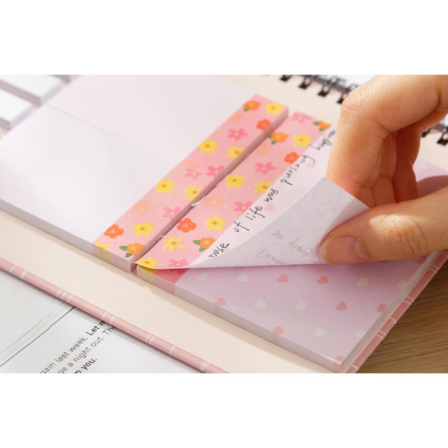 Eine Hand zieht einen floralen Haftnotiz von einem rosa Notizbuch ab und enthüllt eine handgeschriebene Nachricht darunter.