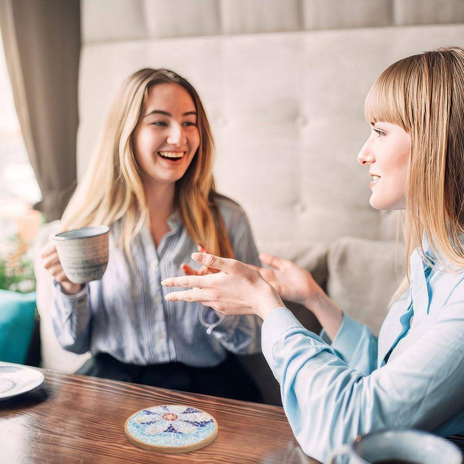 Zwei Frauen unterhalten sich an einem Tisch in einem Café, lächeln und halten Getränke in der Hand, während eine dekorative Untersetzer auf dem Tisch sichtbar ist.