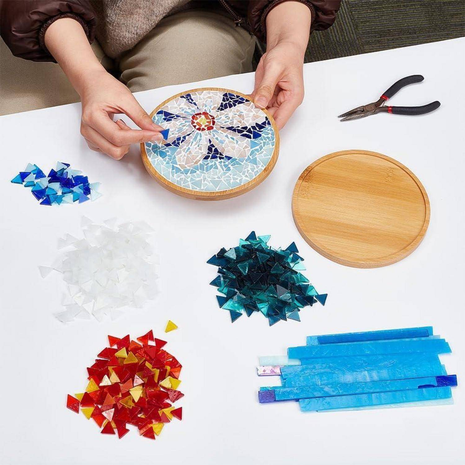 Person, die blaue Mosaikfliesen auf einem runden Holztablett anordnet, mit farbigen Fliesenstapeln, einer Zange und einem zusätzlichen Tablett in der Nähe, zeigt ein laufendes Mosaikhandwerk.
