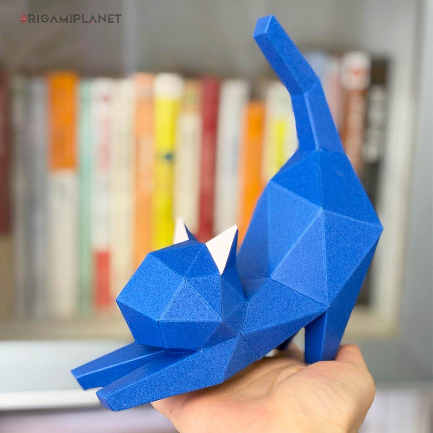 Eine blaue geometrische Papierkatzenfigur sitzt auf der Hand einer Person, während ein unscharfes Bücherregal im Hintergrund zu sehen ist.