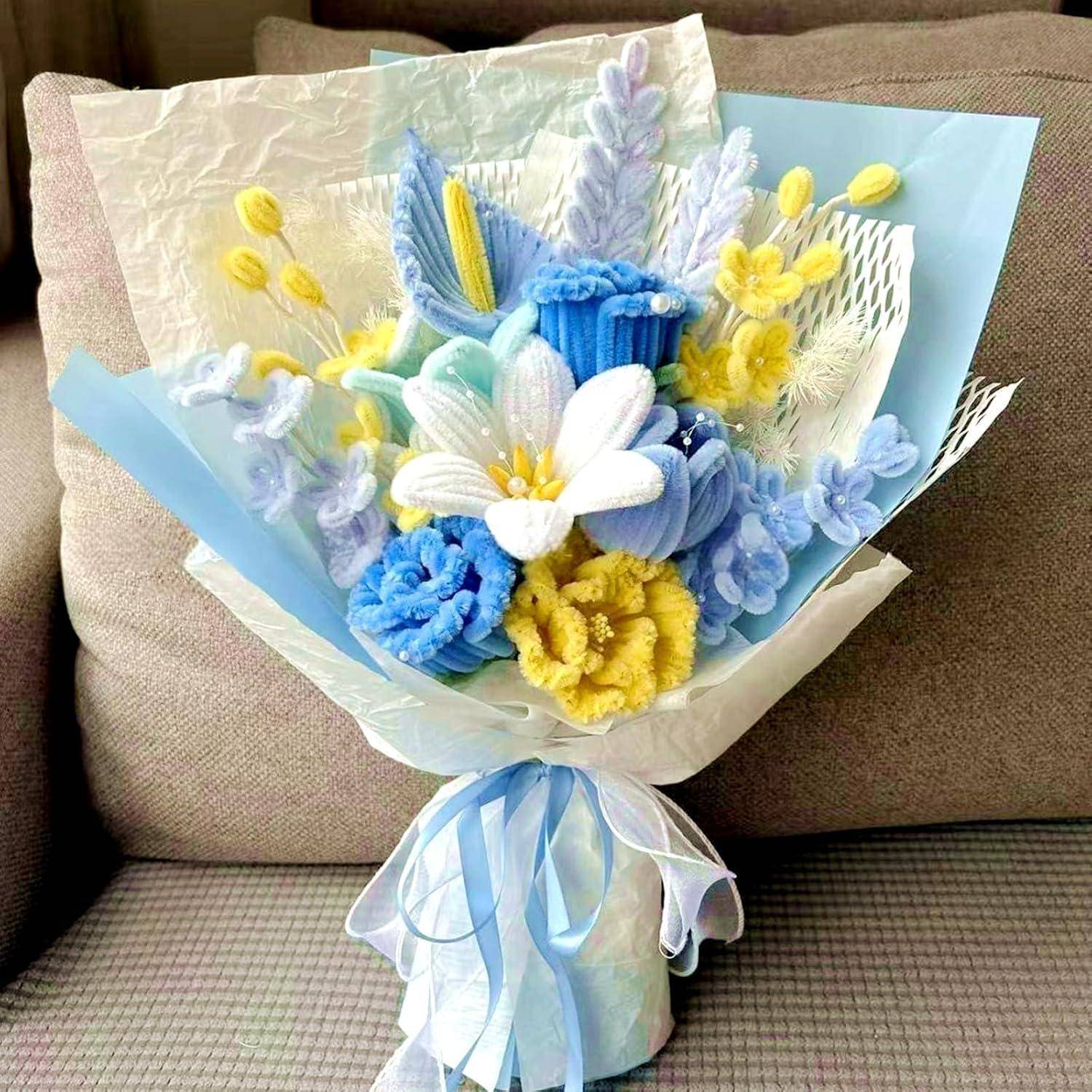 Ein zartes Bouquet aus gestrickten und Papierblumen in Blau, Gelb und Weiß, eingewickelt in durchsichtiges blaues Papier mit einer Schleife.
