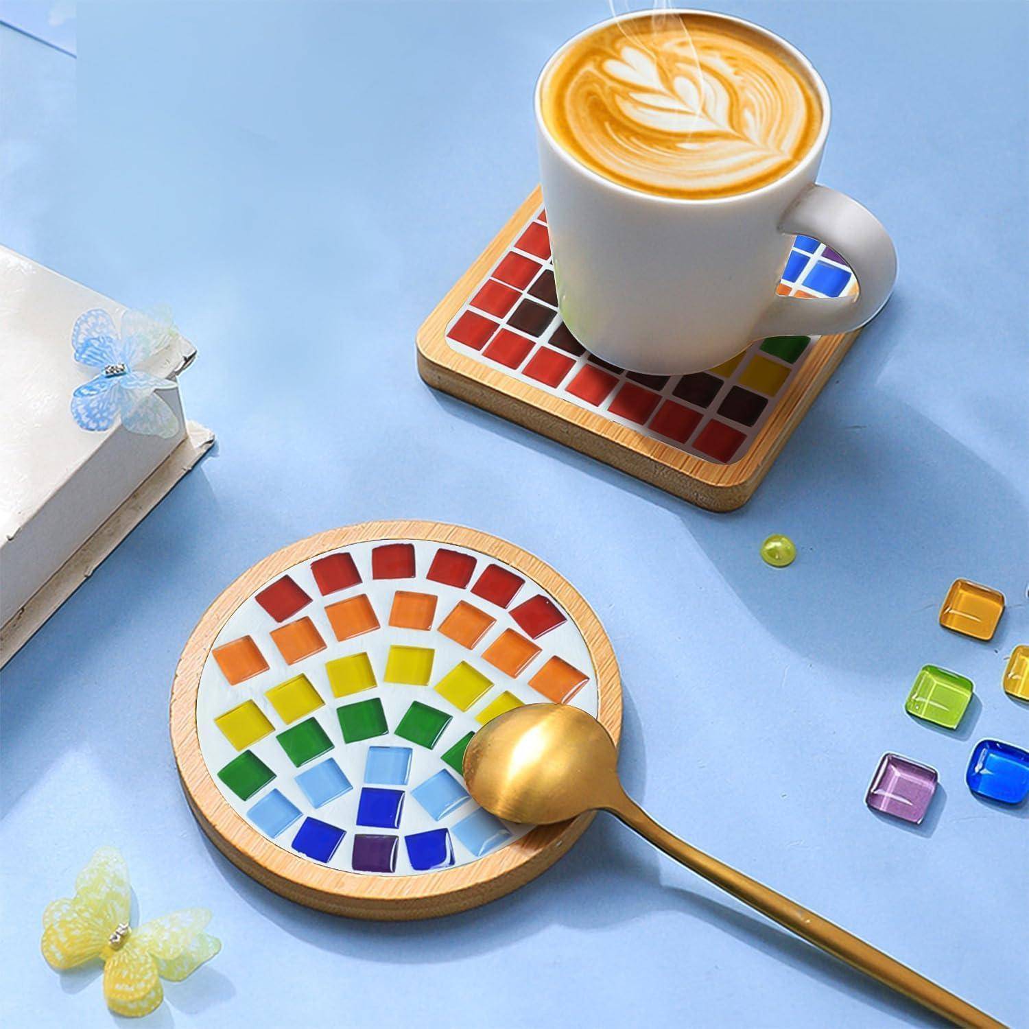 Latte in einer weißen Tasse auf einem Mosaik-Untersetzer, mit einem goldfarbenen Löffel neben einem regenbogenfarbig gekachelten Tablett auf hellblauem Hintergrund.