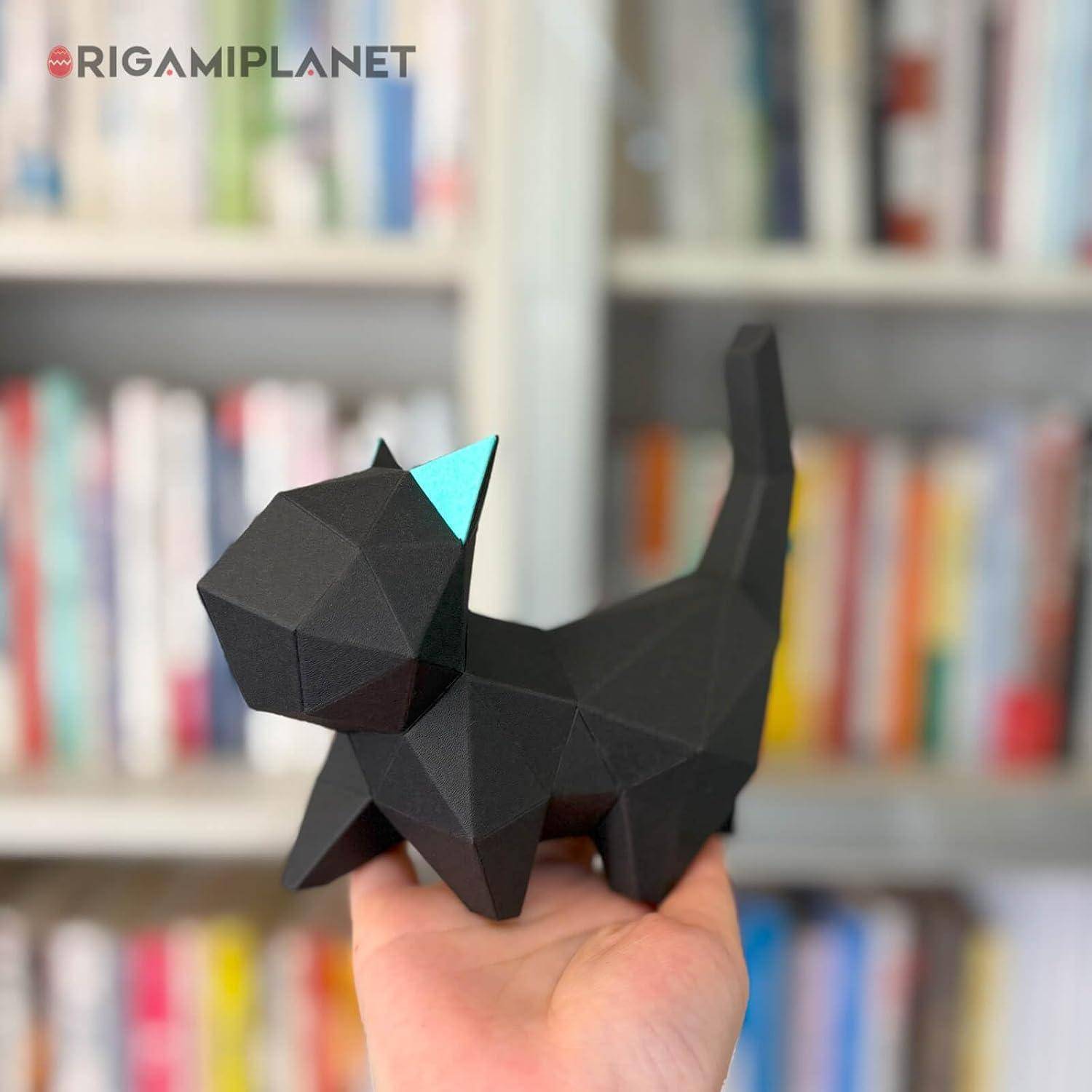 Eine Hand hält ein kleines schwarzes geometrisches Papierkatzenmaterial mit einem türkisfarbenen Ohr vor einem unscharfen Bücherregalshintergrund.