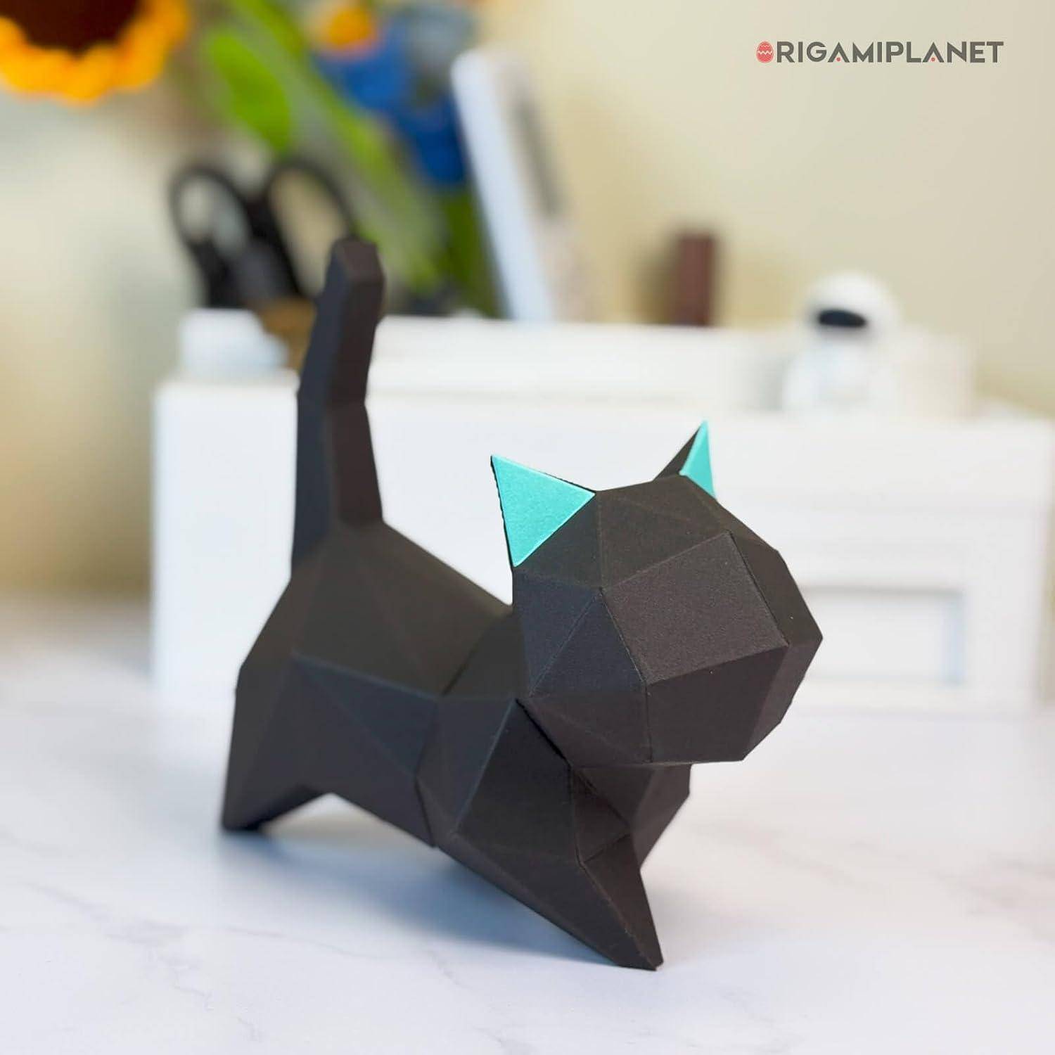 Eine kleine geometrische schwarze Papierkatze mit türkisfarbenen Ohren steht auf einer weißen Oberfläche vor einem unscharfen Innenraum-Hintergrund.