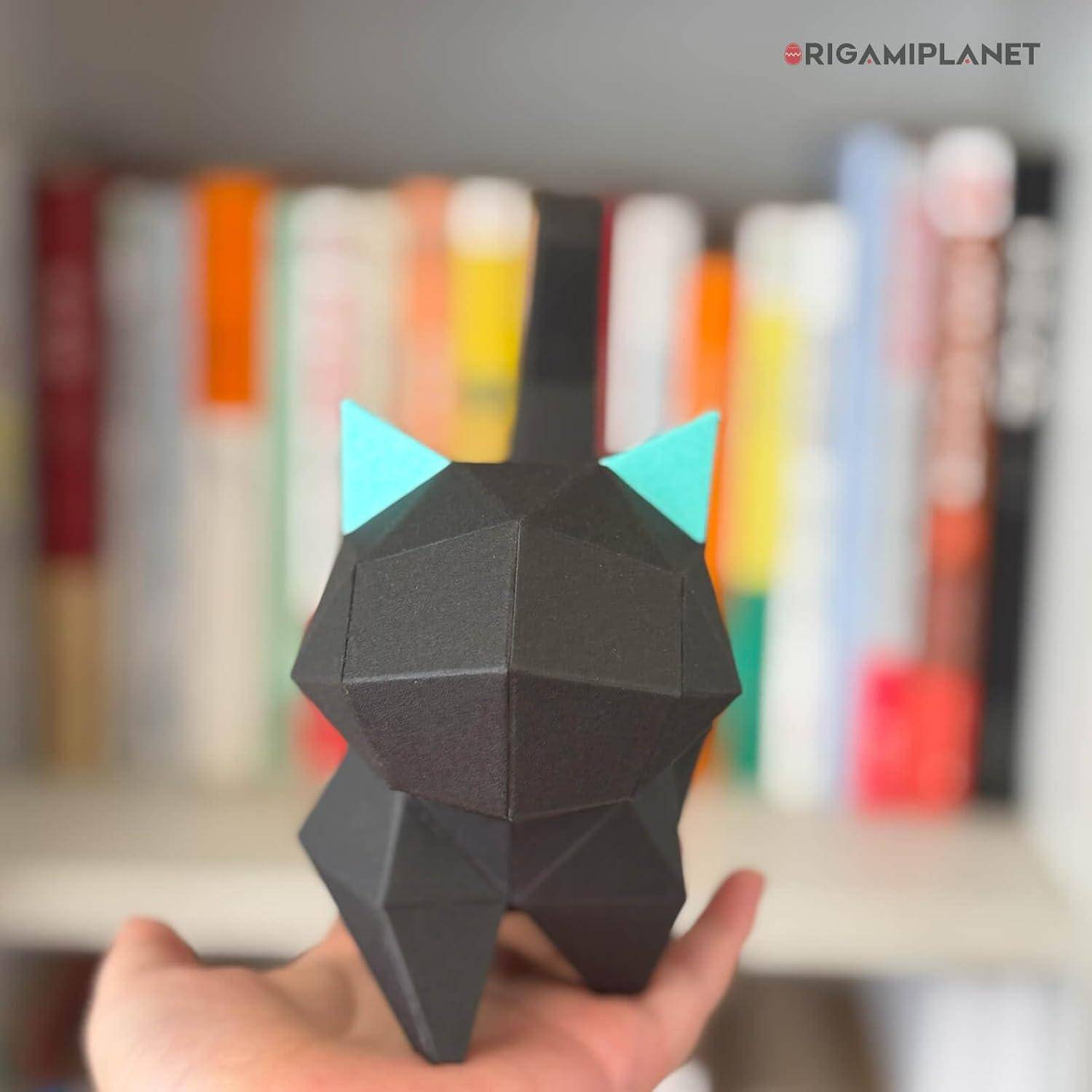 Eine Hand hält eine kleine schwarze geometrische Katzenfigur mit türkisfarbenen Ohren vor unscharfen Büchern auf einem Regal.