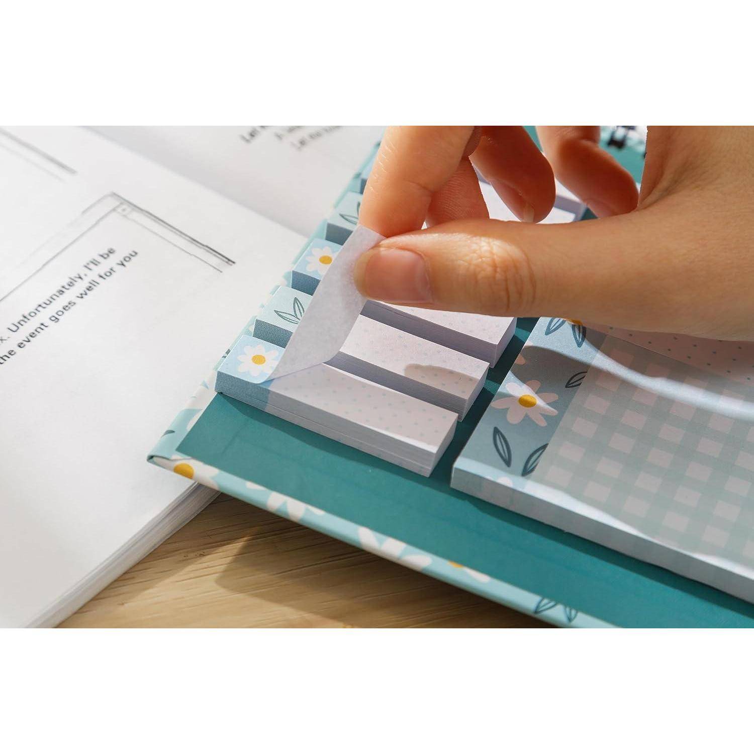 Hand, die eine Papierlasche auf einem Notizbuch oder Scrapbook mit Blumenmuster und gestapelten Abschnitten auf einem Schreibtisch zurückzieht, was auf ein Seiten-Umblätter- oder Organizer-Design hindeutet.