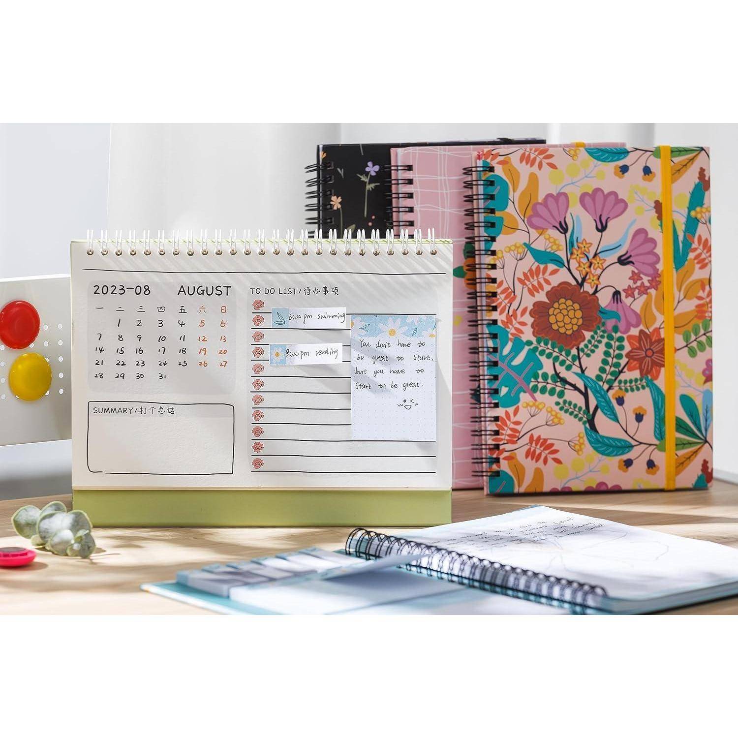 Offene Schreibtisch-Anordnung mit drei spiralgebundenen Notizbüchern; vorderer Planner zeigt einen August-2023-Kalender, eine To-Do-Liste und Zusammenfassungsfelder neben einem bunten blumigen Notizbuch.