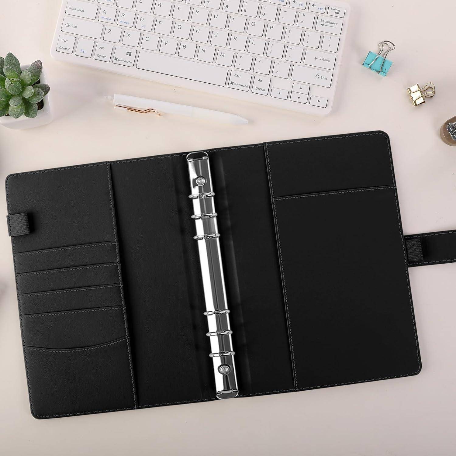Offener schwarzer Ring-Bindeplaner mit mehreren Taschen liegt auf einem Schreibtisch neben einer Tastatur, einem Stift, einer Pflanze und Aktenklammern.