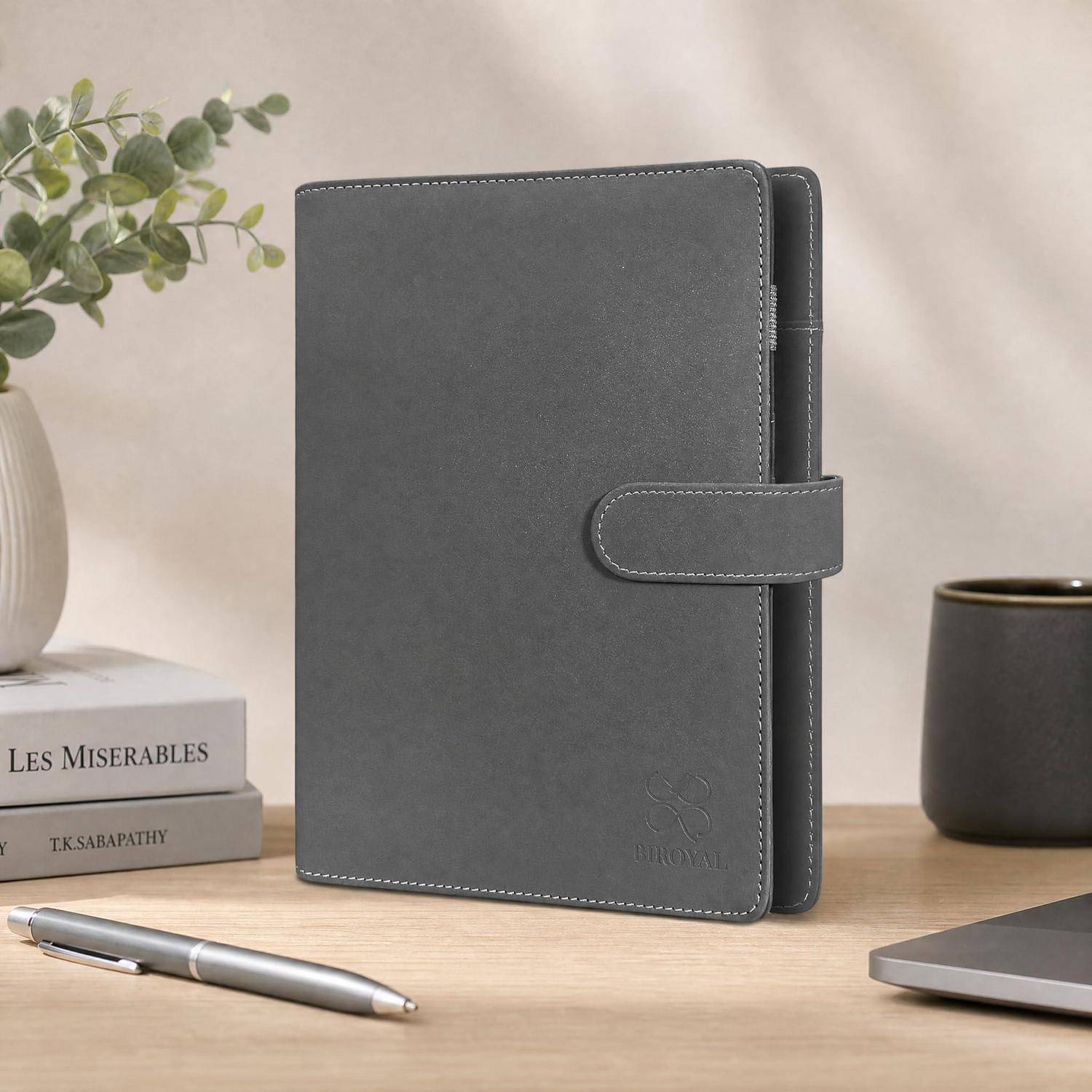 Grauer Ledereinband oder Planer, der auf einem Schreibtisch neben einem Stift, Büchern, einer Tasse und der Ecke eines Laptops steht.