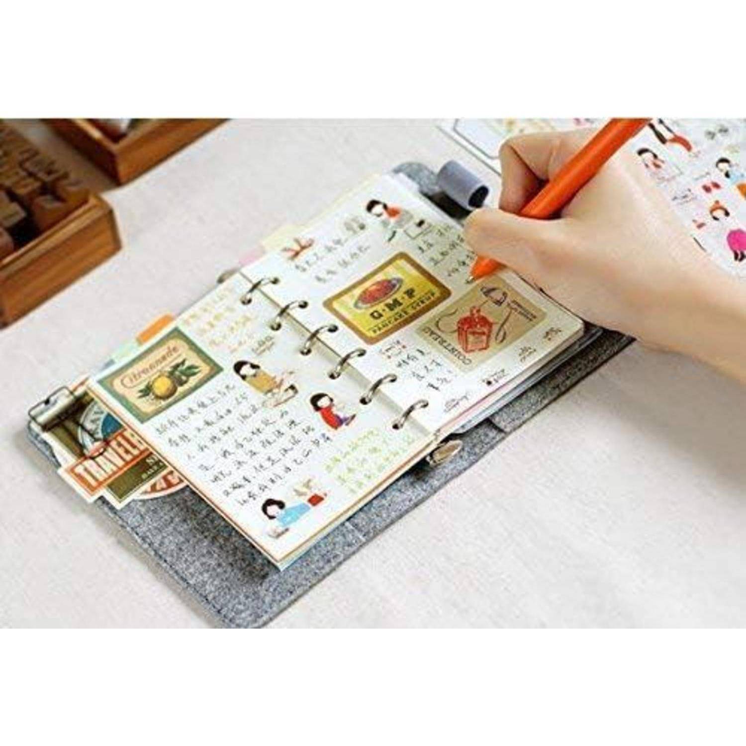 Handschrift in einem kleinen offenen Notizbuch-Planer mit dekorativen Aufklebern, das auf einer grauen Schreibunterlage liegt, während eine Hand mit einem orangefarbenen Stift schreibt.