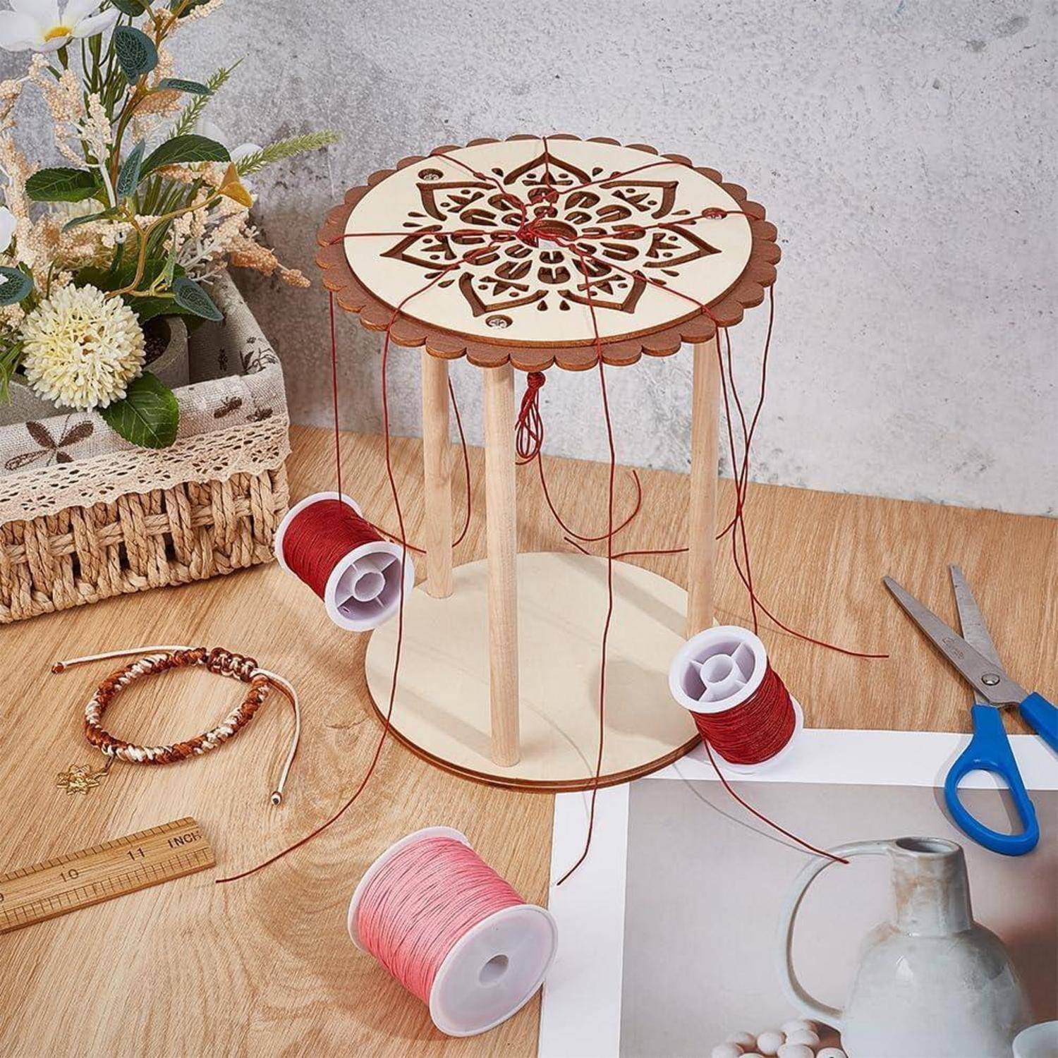 Holzfadenspulenorganizer mit ausgeschnittenem Blumenmuster oben, auf dem rote und rosa Spulen auf einem Basteltisch neben Schere, Lineal und Armbandmaterialien liegen.