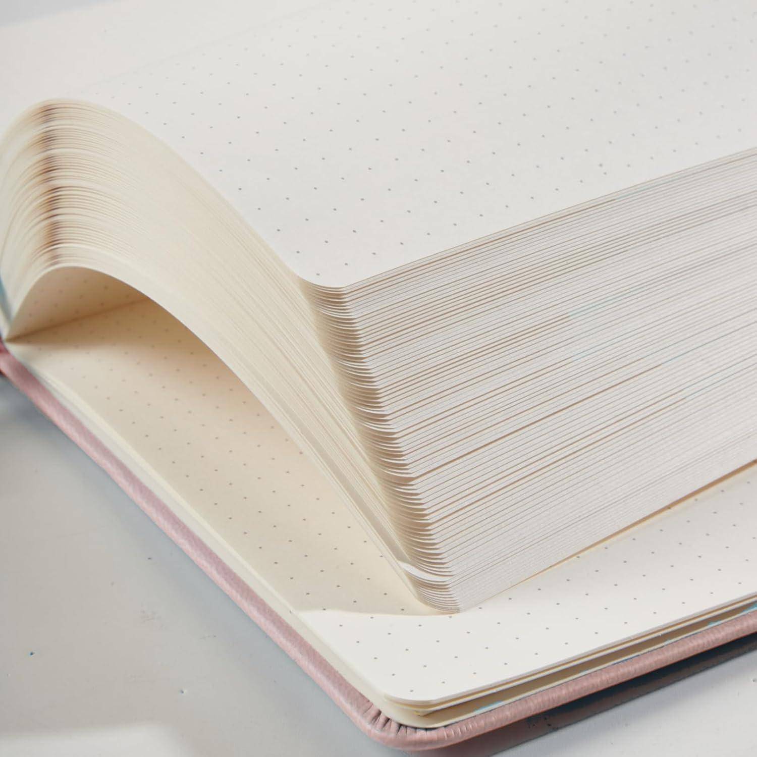 Offenes Punktraster-Notizbuch mit dicken Seiten, die nach oben gebogen sind, zeigt rosafarbig eingefassten Einband und cremefarbenes Papier auf einer hellen Oberfläche.