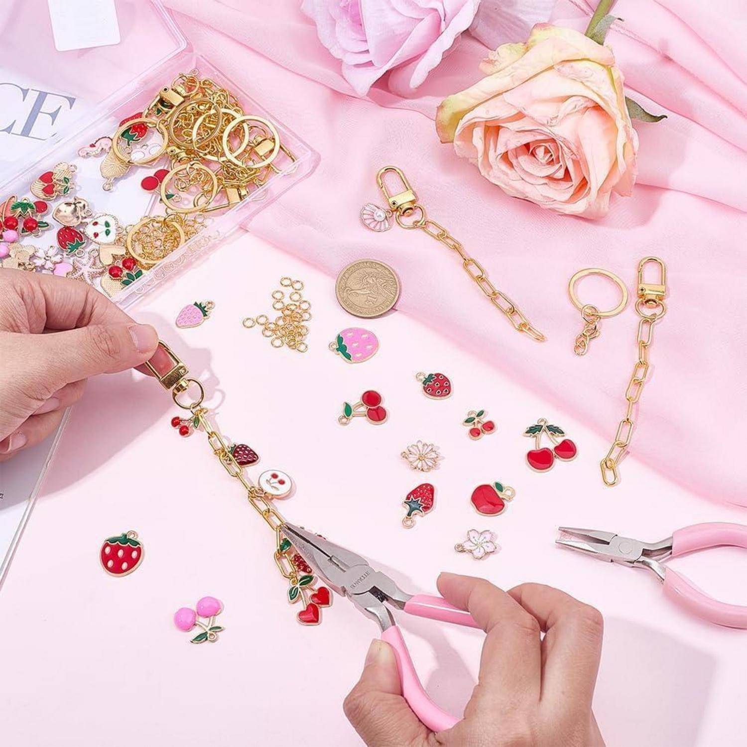 Hände montieren Goldketten-Schlüsselanhänger mit Erdbeer- und Kirsch-Charms auf einer rosa Tischplatte, umgeben von Schmuckzubehör und Zangen.
