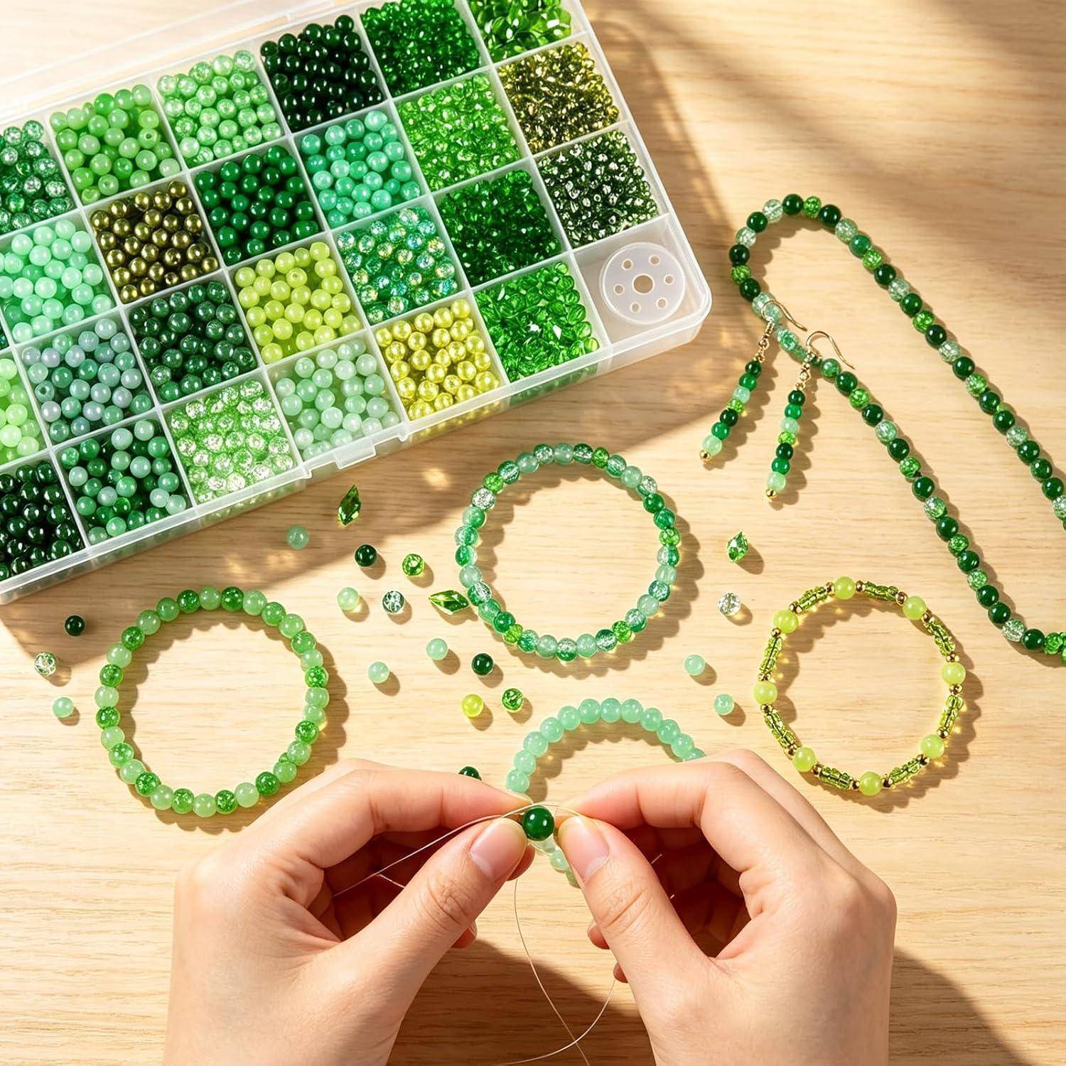 Hände, die grüne Perlen auf eine durchsichtige Schnur fädeln, um Armbänder herzustellen, mit einer Perlenorganisierbox und mehreren fertigen grünen Perlenbändern auf einem Holztisch.