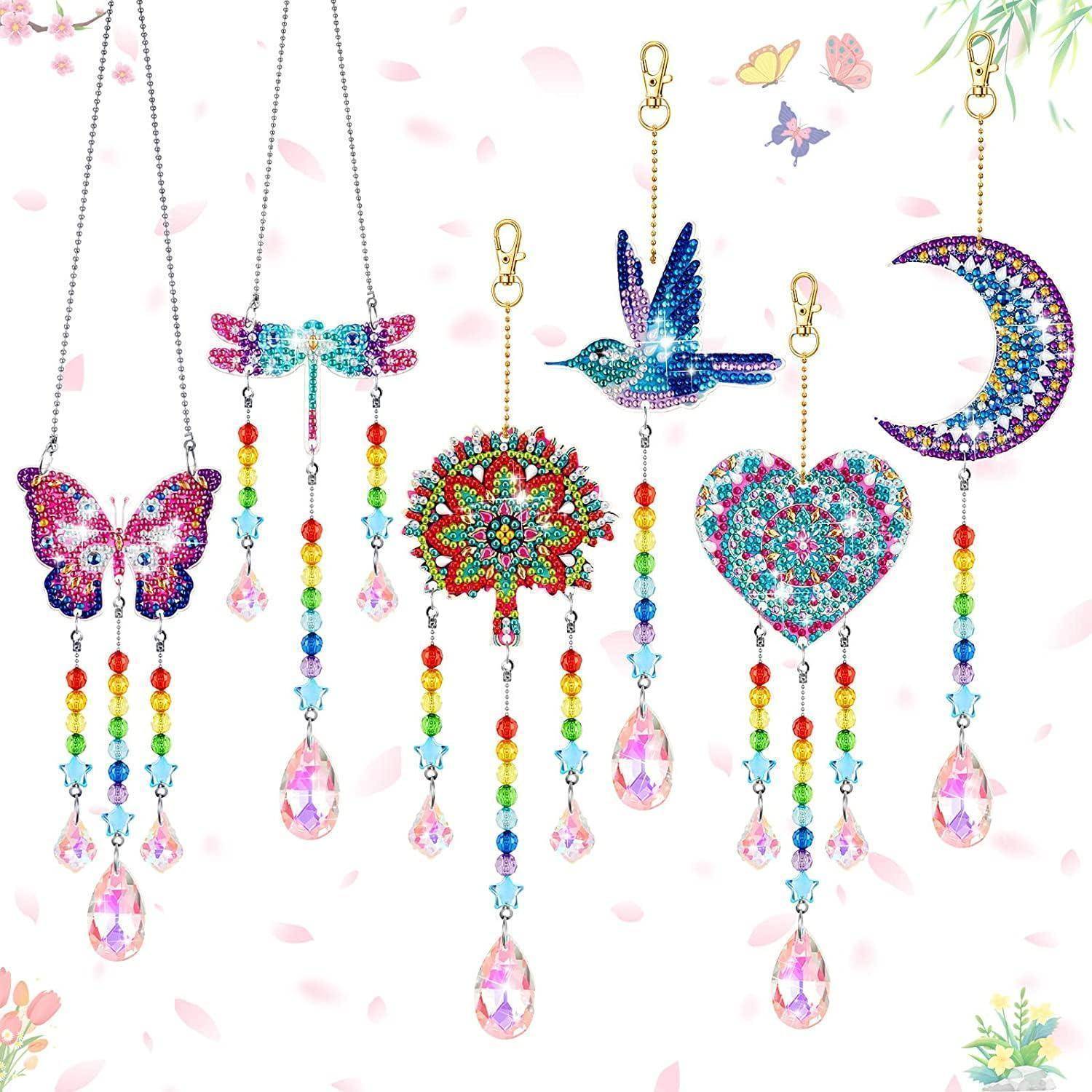 Fünf bunte hängende Kristall-Sonnenfänger – Schmetterling, Libelle, Mandala, Vogel, Herz und Halbmond – auf einem hellen floralen Hintergrund.