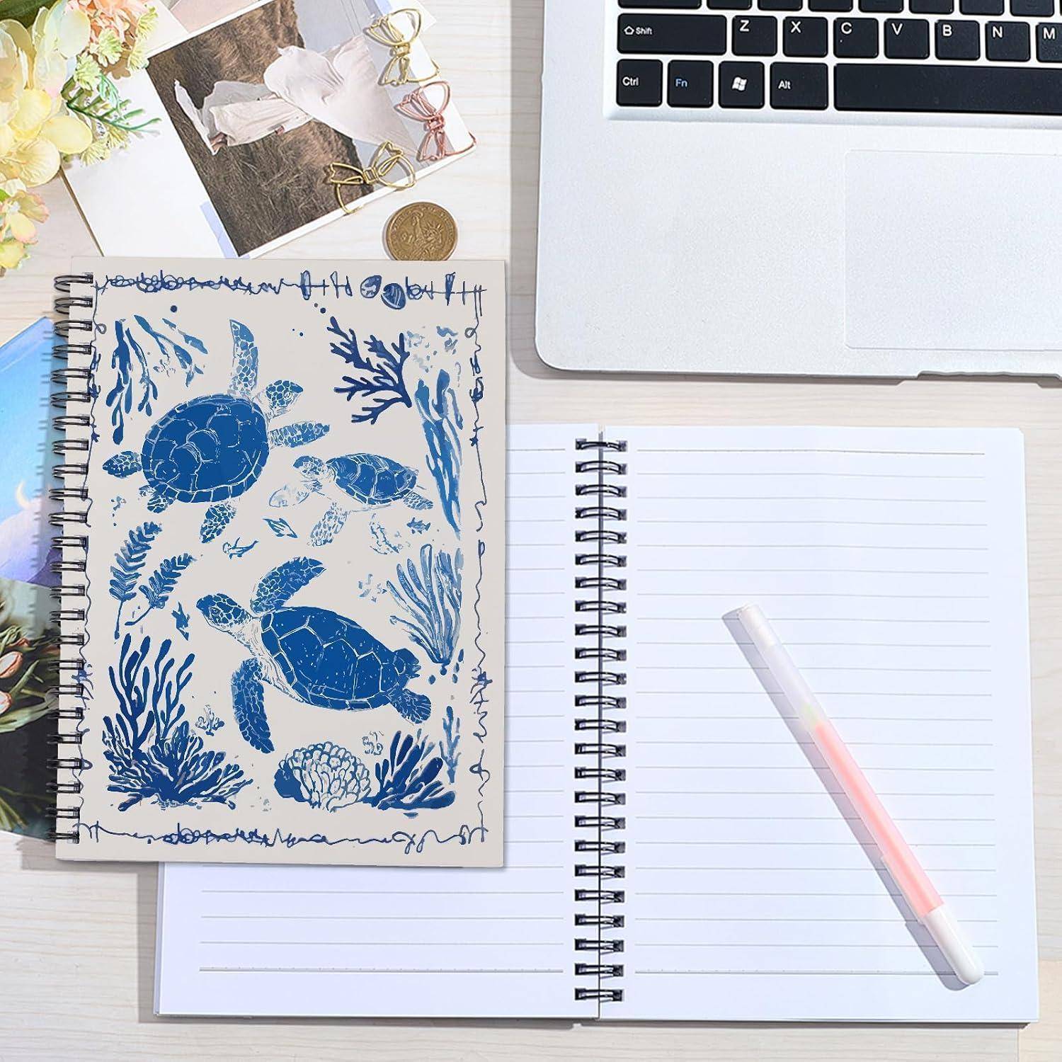 Draufsicht auf einen Schreibtisch mit einem offenen, leeren Laptop, zwei Spiralblöcken, einem Stift und einem Skizzenbuch mit einer blauen Meeresschildkröten-Illustration, was auf einen kreativen Arbeitsplatz hindeutet.