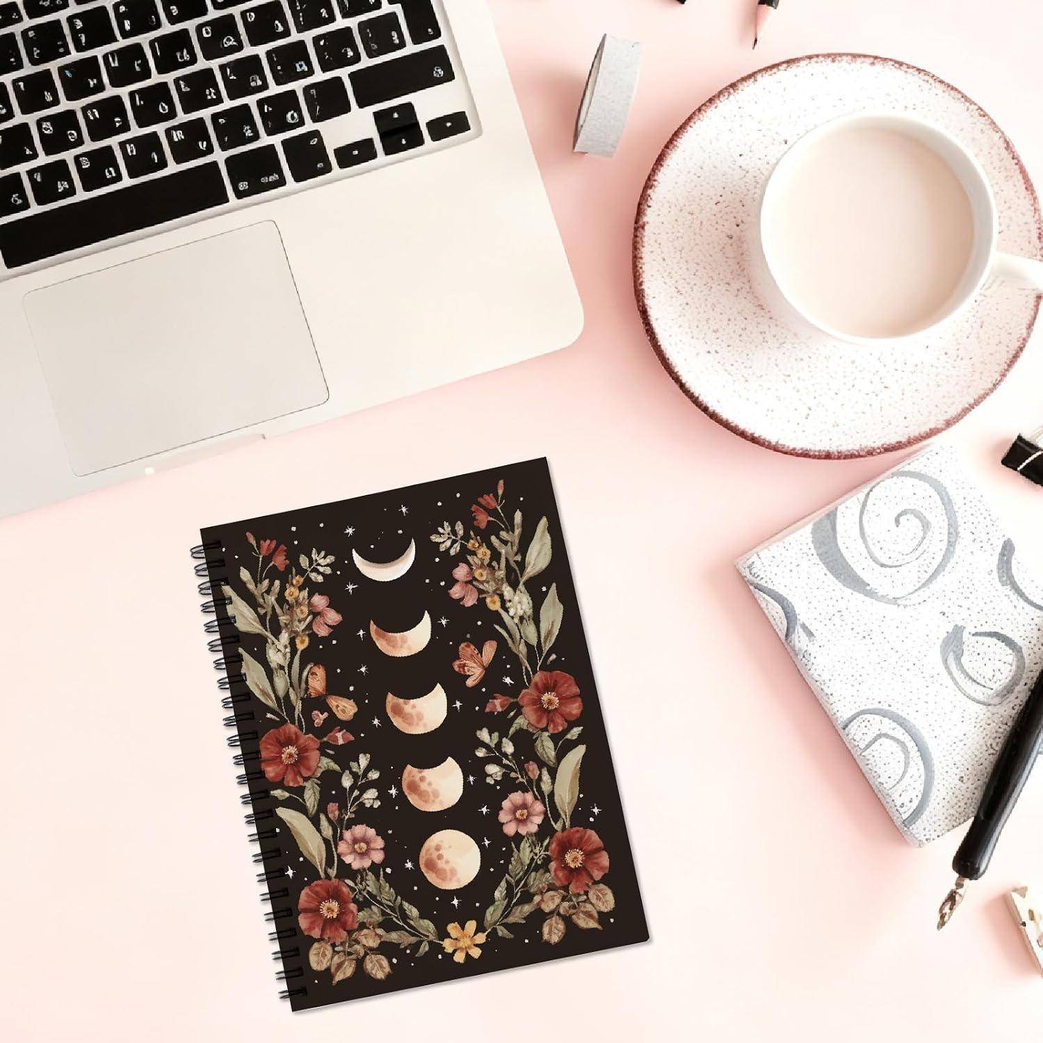 Schreibtisch-Flatlay mit einem Laptop, einer Kaffeetasse, einem gemusterten Notizbuch, einem Stift und einem gefalteten Tuch auf einer rosa Oberfläche.