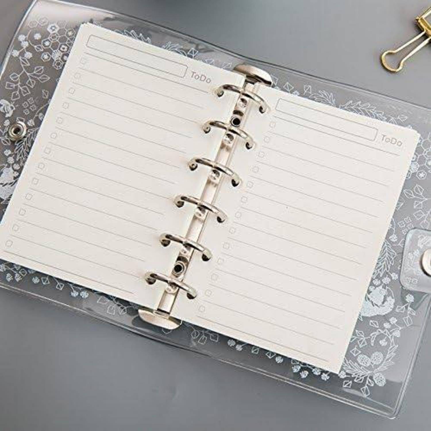 Offenes ringgebundenes To-Do-Notizbuch mit leeren linierten Seiten auf einem grauen Schreibtisch, gezeigt in einem klaren dekorativen Umschlag.