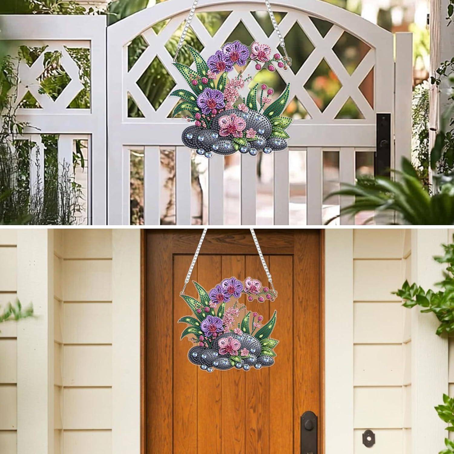 Ein dekoratives Blumen-Wandbehang mit rosa und violetten Blüten, grünen Blättern und grauen Steinen, ausgestellt auf einem weißen Tor und dann an einer Haustür aus Holz.