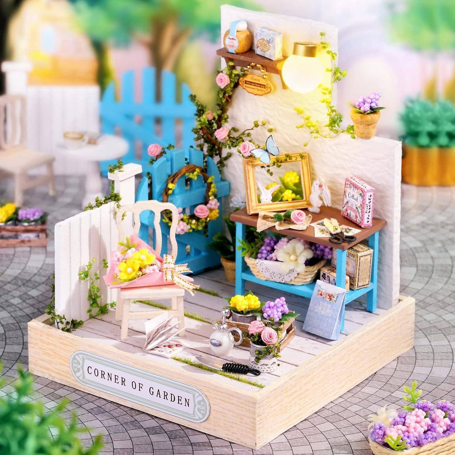 Miniatur-Gartenbastlszene mit einem weißen Stuhl, blauem Tor, Werkbank, Lampe, Blumen und einem Schild mit der Aufschrift „ECKE DES GARTENS