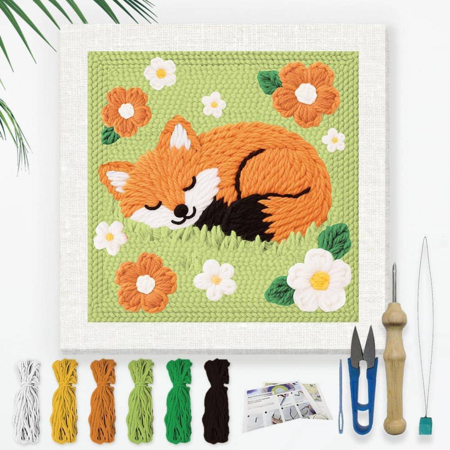 Gestickte Darstellung eines schlafenden orangefarbenen Fuchses, umgeben von Blumen, gezeigt mit Garn, Werkzeugen und einer Anleitung für ein Handwerksprojekt.