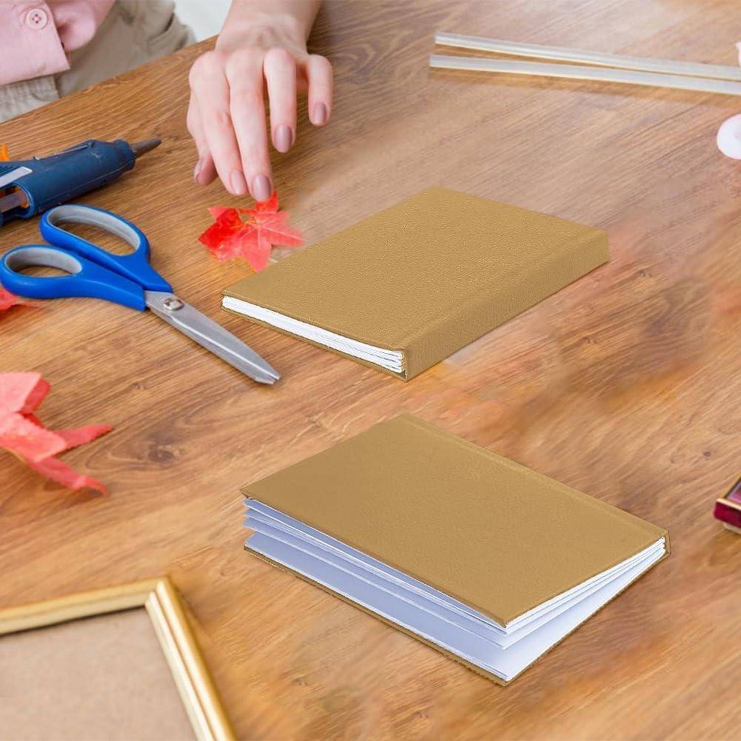 Person, die handgefertigte Bücher auf einem Holztisch anfertigt, mit Schere, Heißklebepistole, dekorativem Papier und zwei in Arbeit befindlichen gebundenen Notizbüchern.