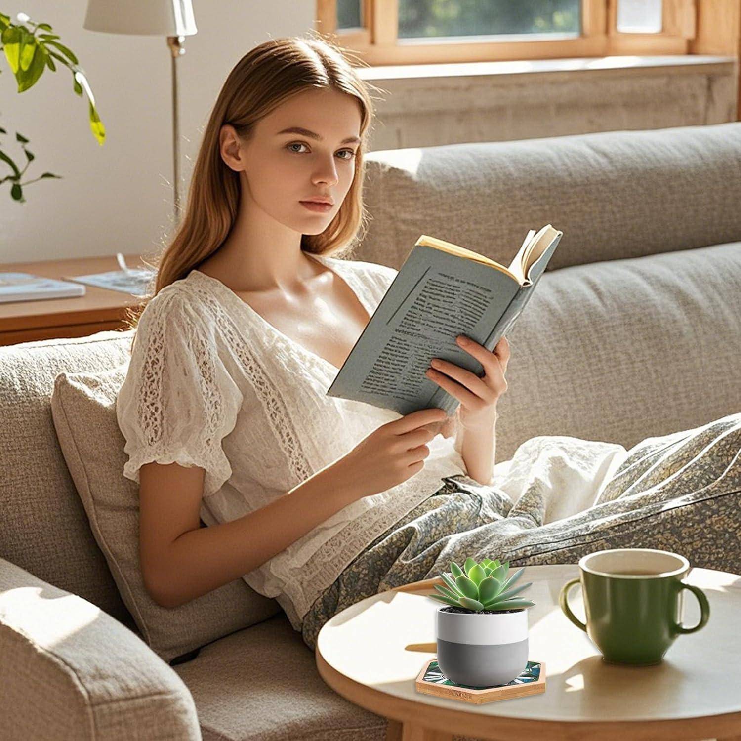Junge Frau, die ein Buch auf einem sonnigen Sofa neben einem kleinen Tisch mit einer Topfpflanze und einer grünen Tasse liest.