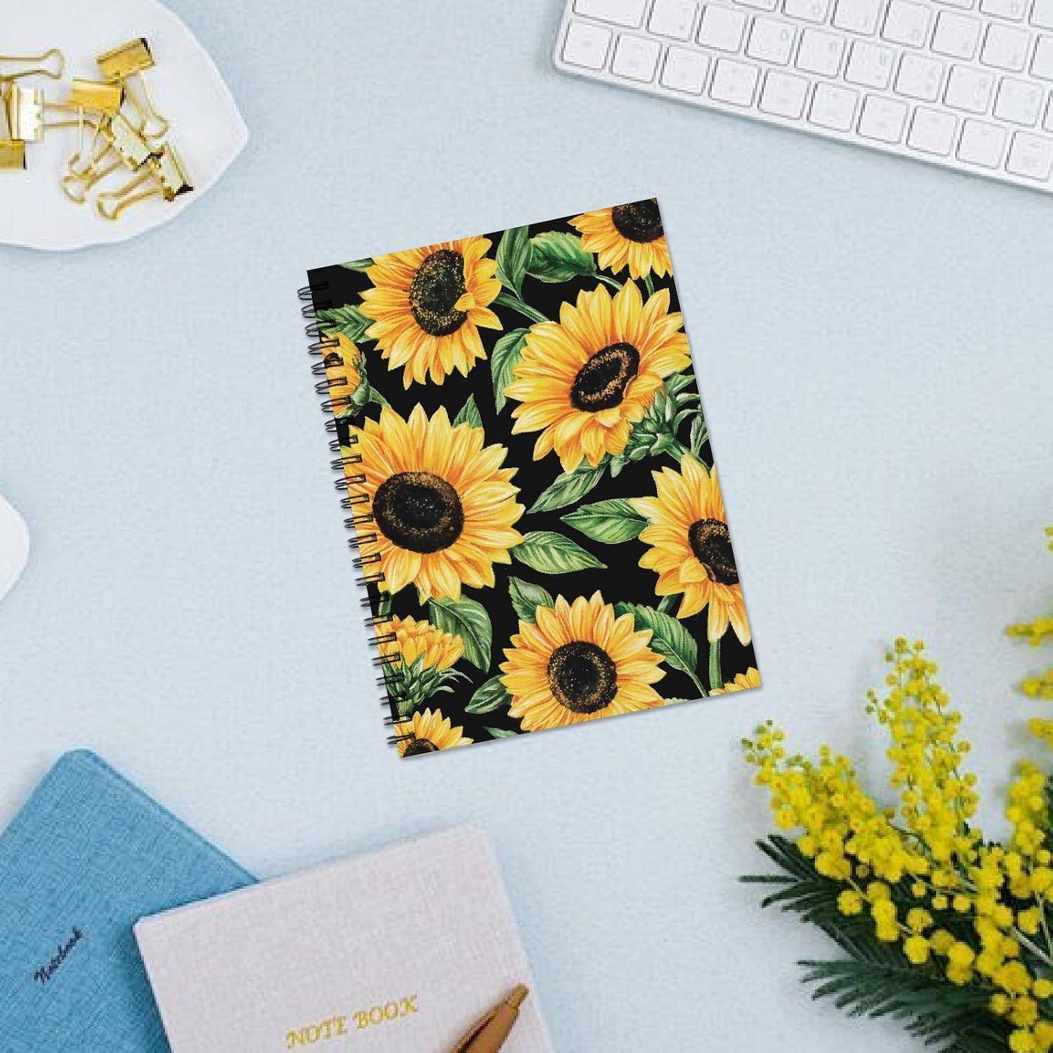 Spiralblock mit Sonnenblumenmuster auf einem hellen Schreibtisch, umgeben von einer Tastatur, einem Band, einem Stoff und gelben Blumen.