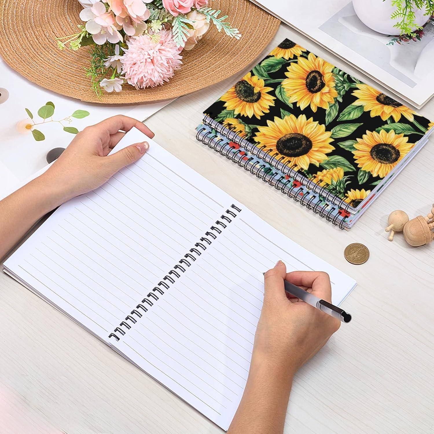Hände schreiben in einem offenen Notizbuch mit Linien auf einem weißen Schreibtisch neben einem Notizbuch mit Sonnenblumenmuster, einer Münze und Blumen.