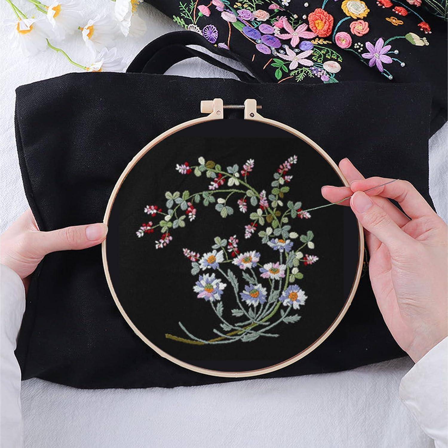 Hände halten einen Stickrahmen mit einem Blumenstichmuster auf schwarzem Stoff, mit bunten fertigen bestickten Blumen im Hintergrund.